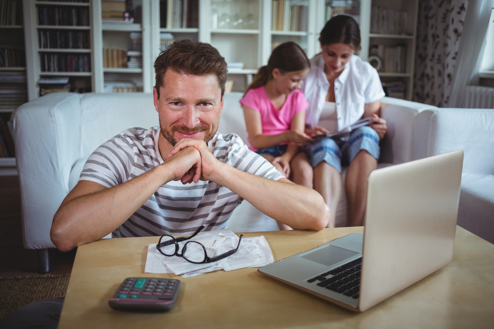 Smiling man sitting at table with bills and laptop while his wife and daughter sitting on sofa